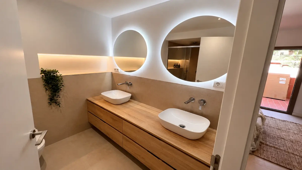 Reforma de baño con mueble de madera de doble seno y espejos circulares retroiluminados en Barcelona